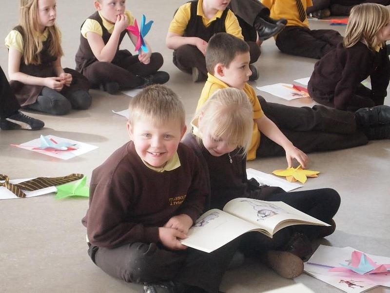 A boy in a school jumper smiles at the camera while sharing a book with a classmate